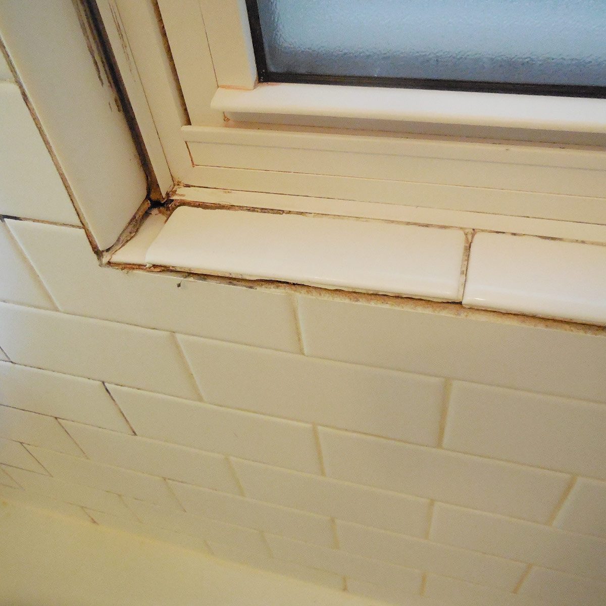 A tiled corner of a bathroom window shows peeling grout and discoloration, indicating potential moisture damage near the edge of the glass pane.