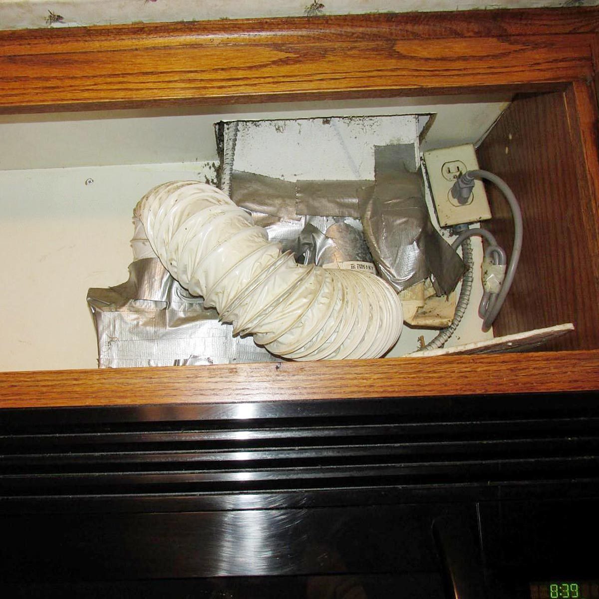 A white flexible duct is attached above a power outlet, surrounded by duct tape and debris, situated in a kitchen cabinet above a black appliance.