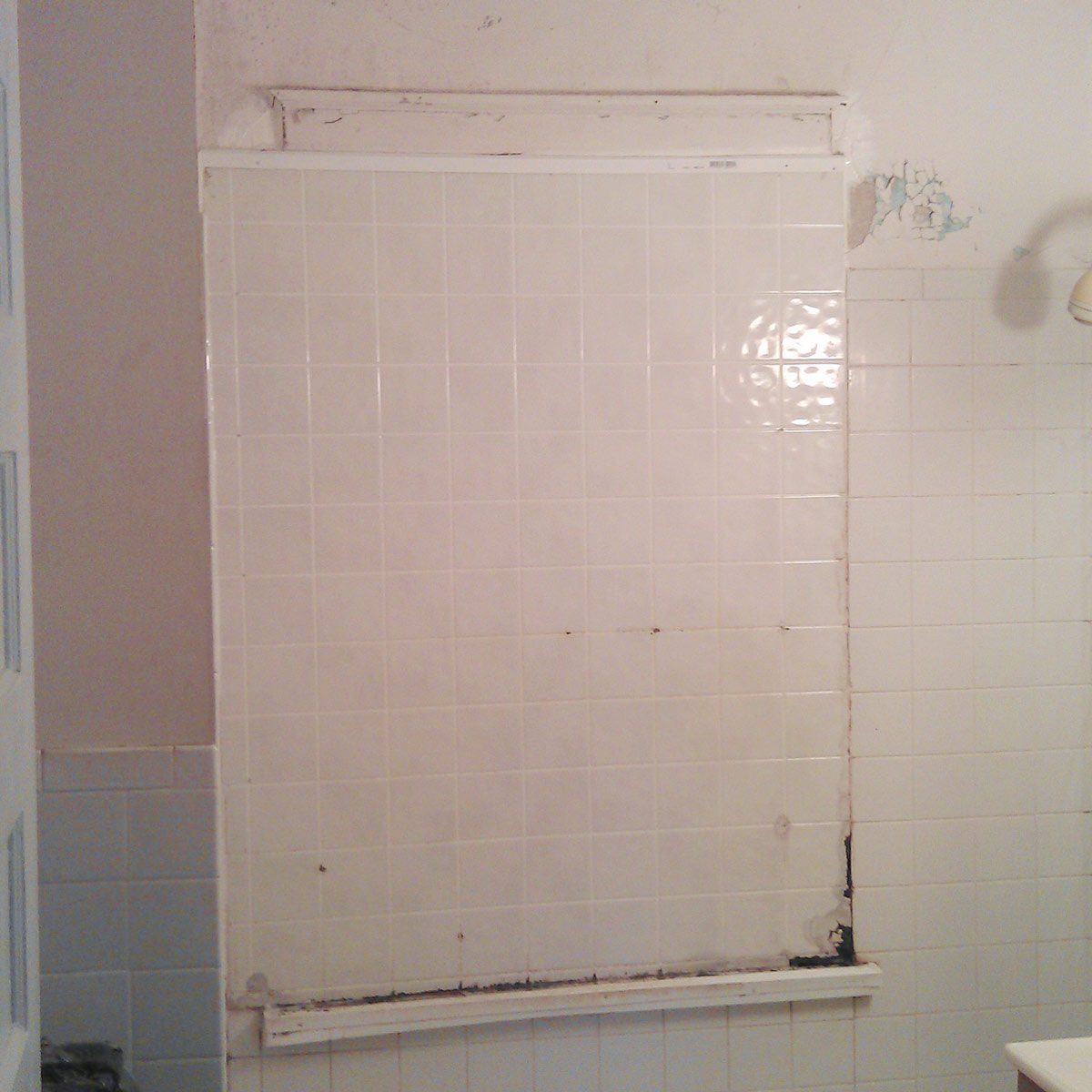 A large, white tiled wall, partially covered, shows signs of wear and peeling edges, situated in a bathroom with faint shower fixtures nearby.