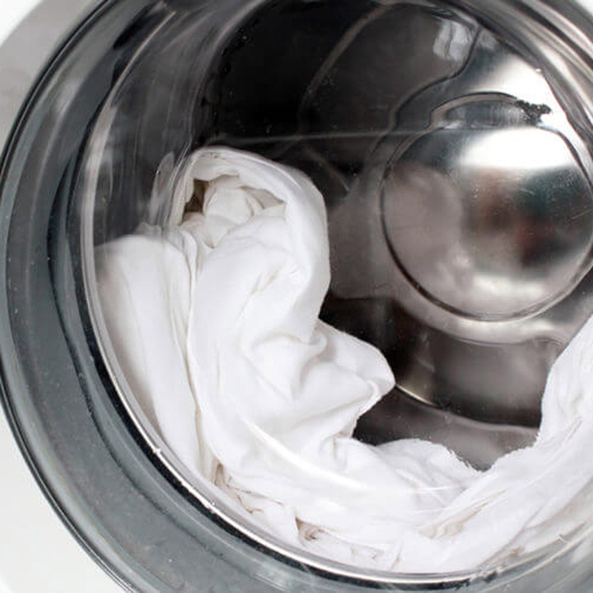 A white fabric is tumbling inside a metallic washing machine drum, surrounded by shiny metal, reflecting the smooth interior surface.