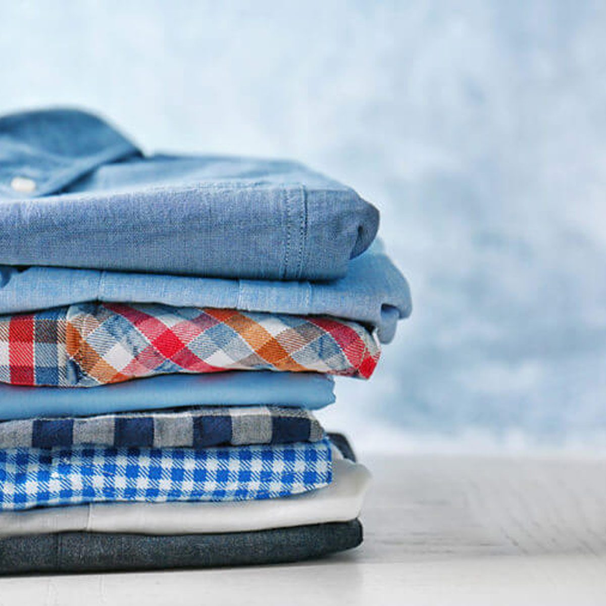Stacked shirts in various colors and patterns are neatly arranged on a table with a soft, blurred blue background.