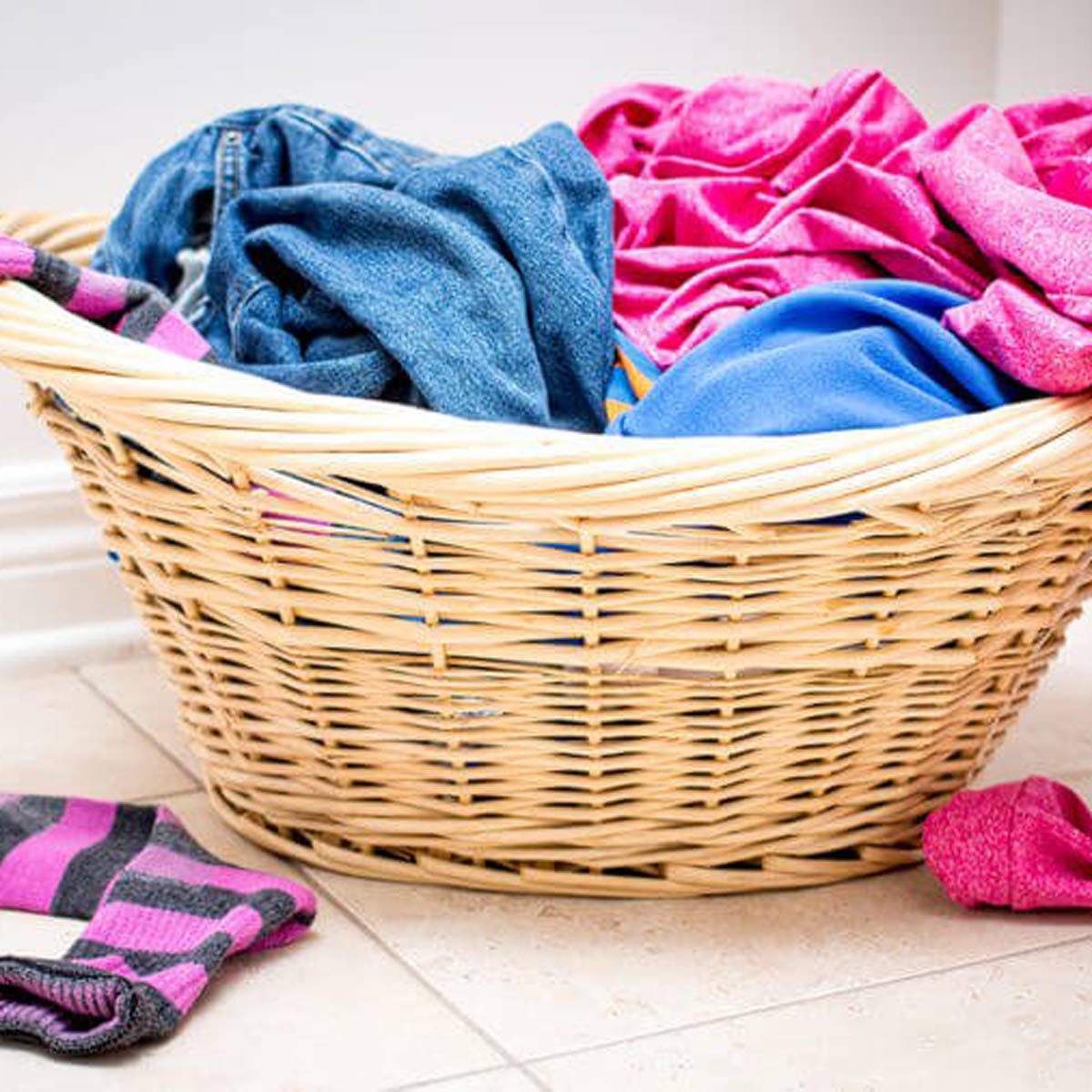 A wicker laundry basket overflows with colorful clothes, including jeans and various fabrics, positioned on a tiled floor in a bright indoor space.
