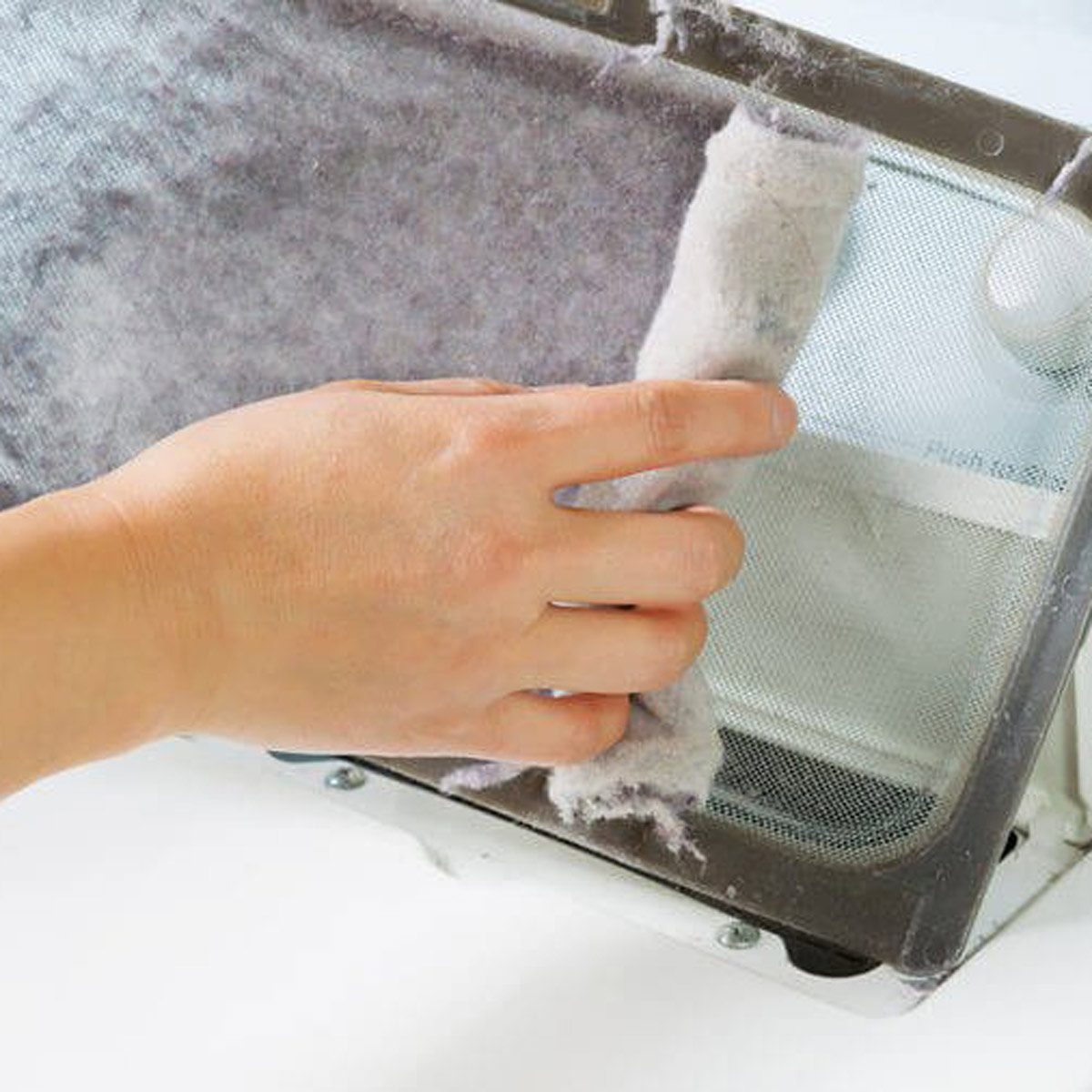 A hand removes lint from a filter, cleaning it near a white appliance, suggesting maintenance of a dryer or another household device.
