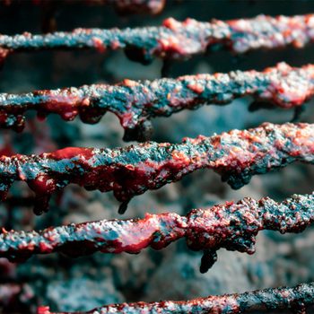 Charred grill grates are dripping with darkened sauce, indicating recent cooking. The smoky background suggests a barbecue or grilling environment.