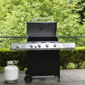 A stainless steel gas grill with an open lid sits on a patio, accompanied by a propane tank, surrounded by green foliage and a fence.