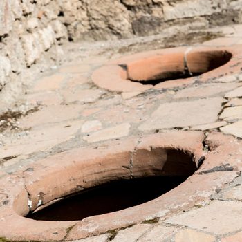 Two round, terracotta openings set into a stone pavement reveal dark interiors, suggesting a historical setting with aged stone walls nearby.