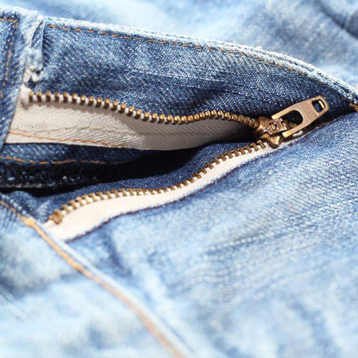 A partially zipped denim zipper reveals its metal teeth, set against a light blue fabric background, showcasing the inner lining of the jeans.
