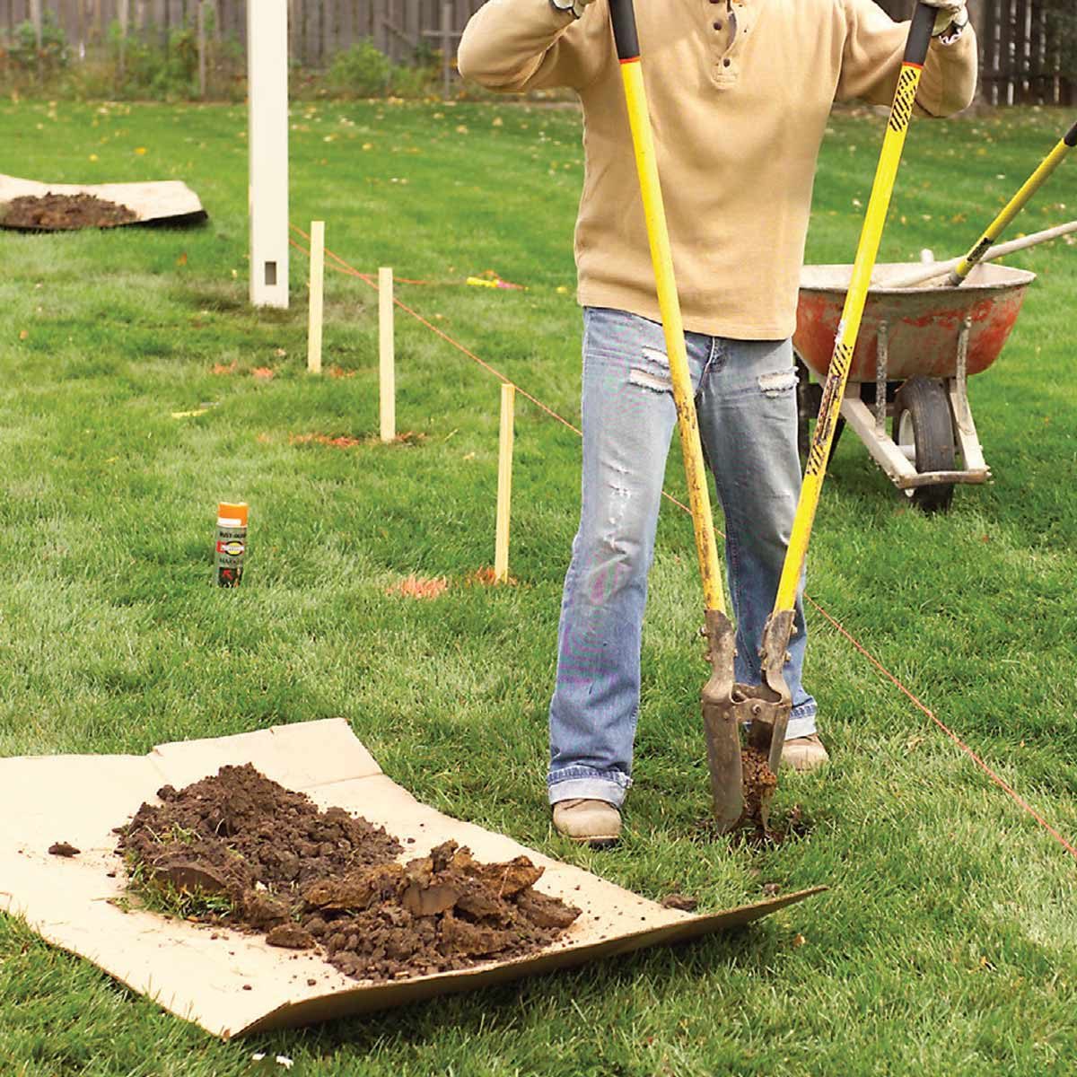 A person uses a post hole digger to remove soil from a grassy area, with a wheelbarrow and a marked construction site in the background.