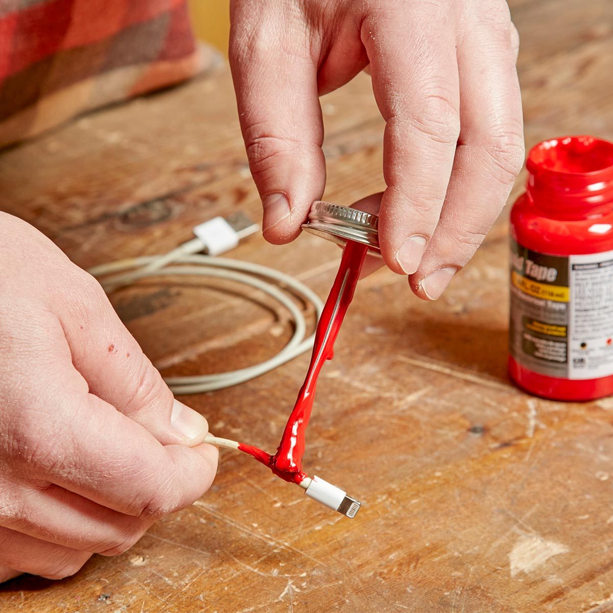 A hand applies red liquid adhesive to the damaged end of a charging cable, with tools and a bottle of adhesive set on a wooden surface.