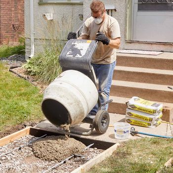 A man pours concrete from a mixer into a prepared area, surrounded by grass, gravel, and concrete mix bags on a sunny day.
