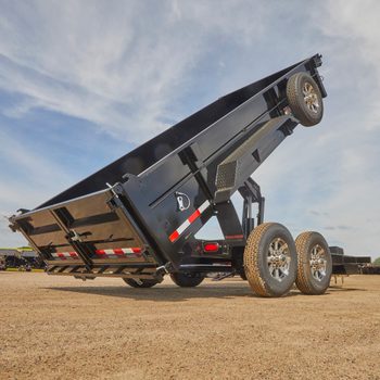 A black dump trailer is lifting its bed, preparing to release a load. It stands on a gravel surface under a clear blue sky.
