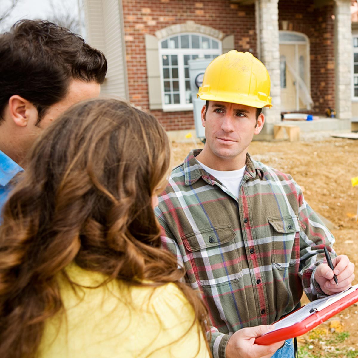 A construction worker in a yellow hard hat discusses plans with two people on a building site, surrounded by dirt and a partially constructed house.