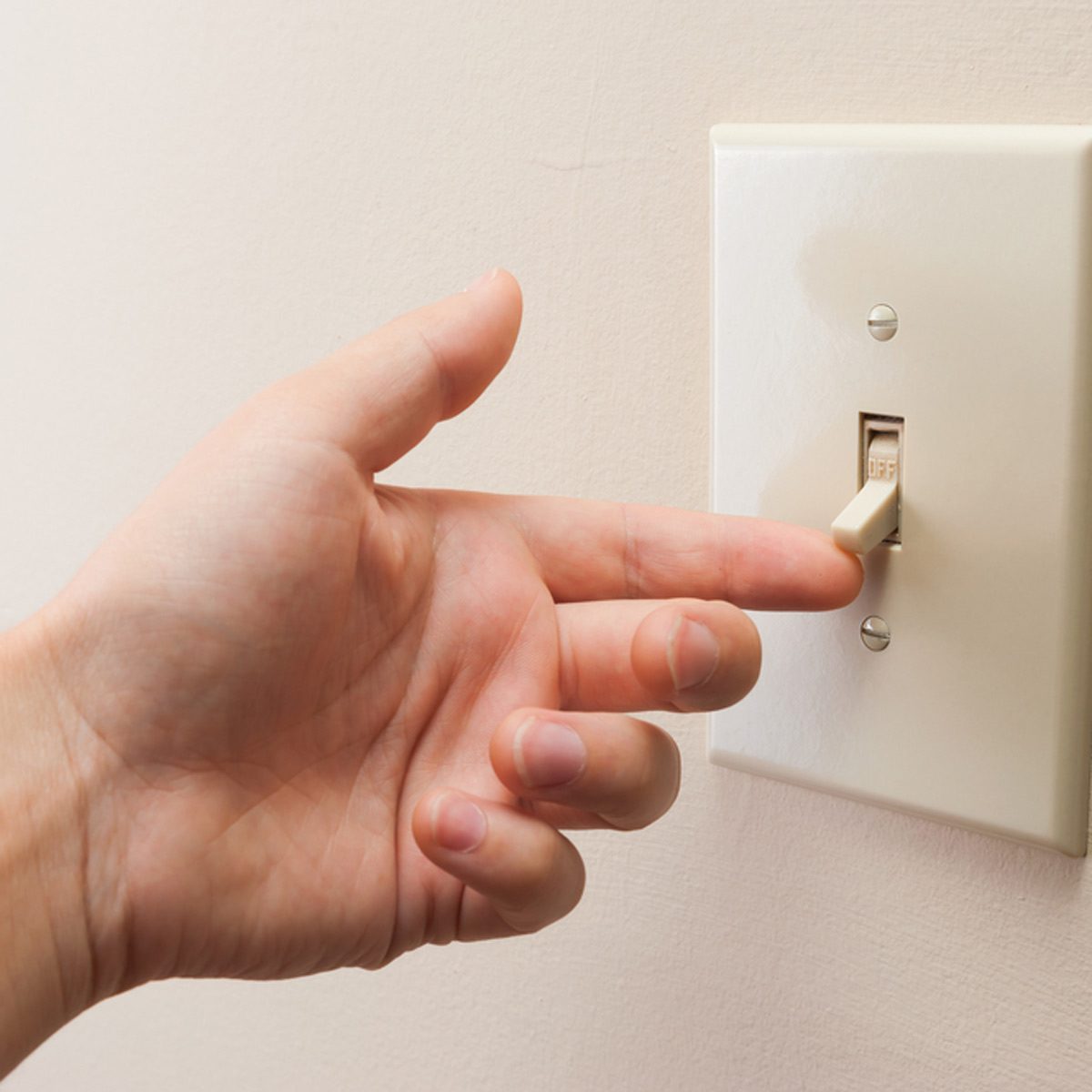A hand reaches towards a light switch, poised to turn it off, against a plain wall background, emphasizing a domestic setting.