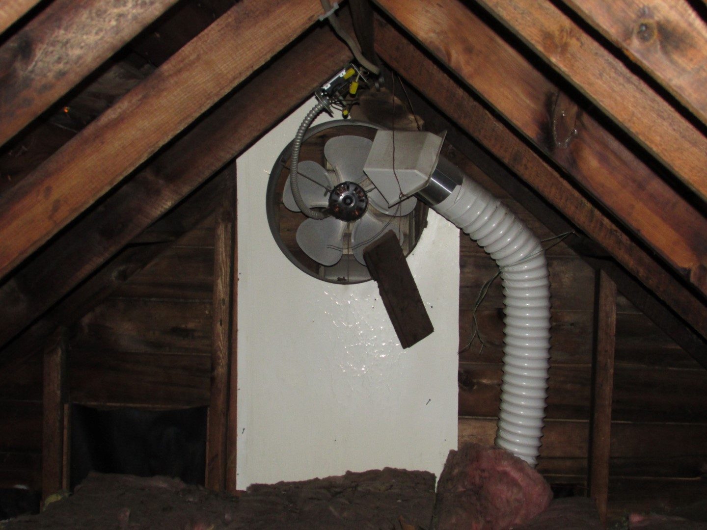 A ventilation fan is mounted on a wall, directing airflow through a flexible duct in a wooden attic, with exposed beams and a gravel floor.