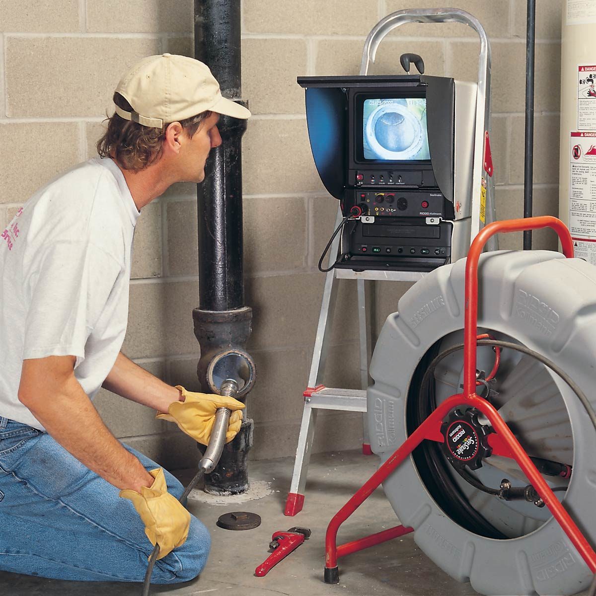 A technician inspects a pipe using a video camera connected to a monitor, positioned beside a large, wheeled reel in a basement setting.
