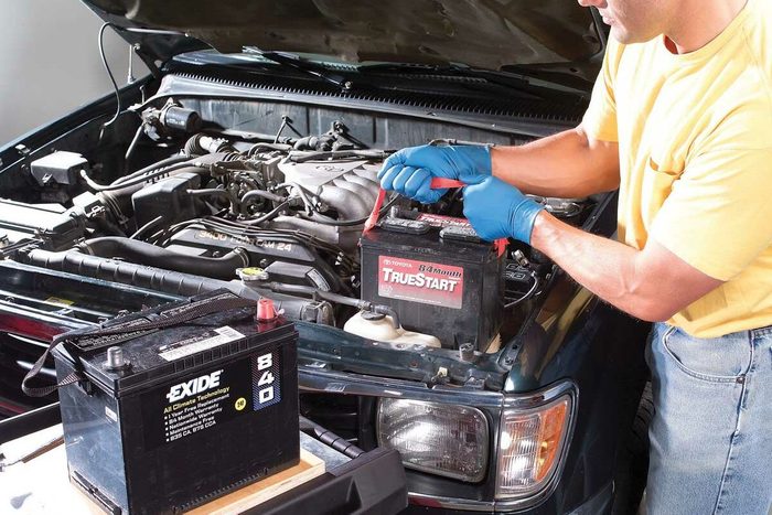 A man replaces a car battery under the hood, using red clips. An EXIDE battery rests on a nearby tool chest in a garage setting.