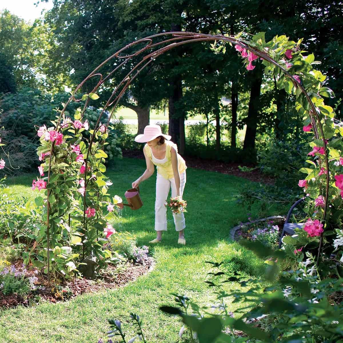 A woman waters flowers under a trellis adorned with pink blooms, surrounded by a lush garden and greenery in bright sunlight.