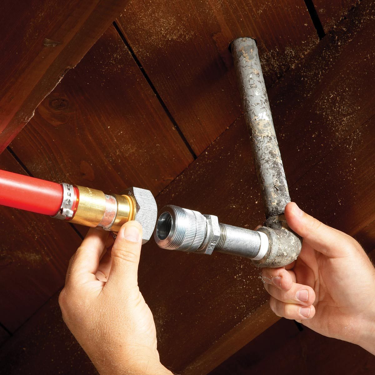 A person connects a red hose to a metal pipe fitting in an overhead structure with wooden beams, indicating plumbing work in progress.