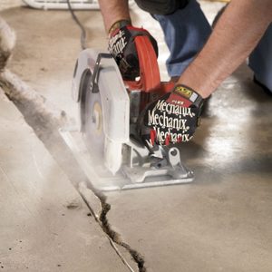 A person uses a circular saw to cut along a crack in a concrete floor, generating dust in a workshop environment.