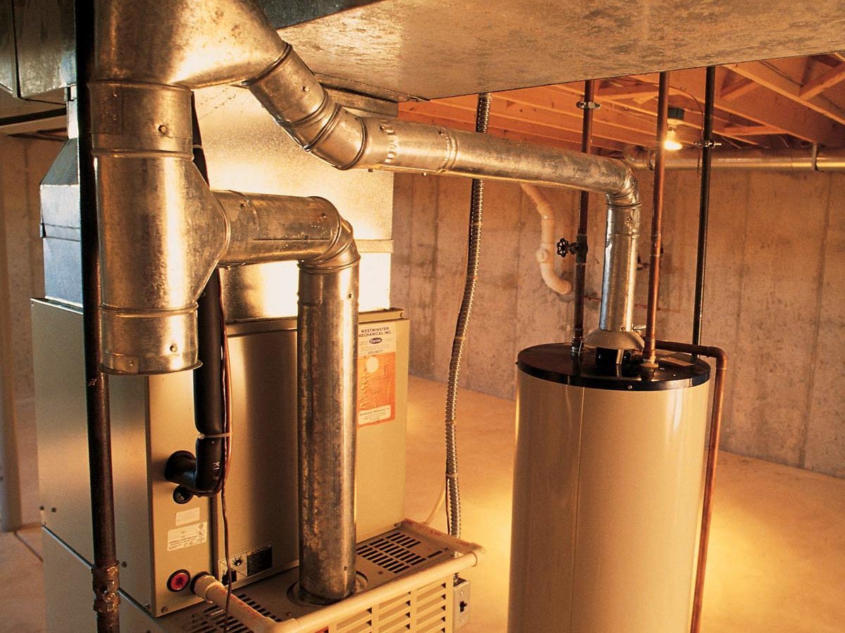 Metal pipes connect a furnace to a water heater in a brightly lit basement with concrete walls, emphasizing domestic utility infrastructure.