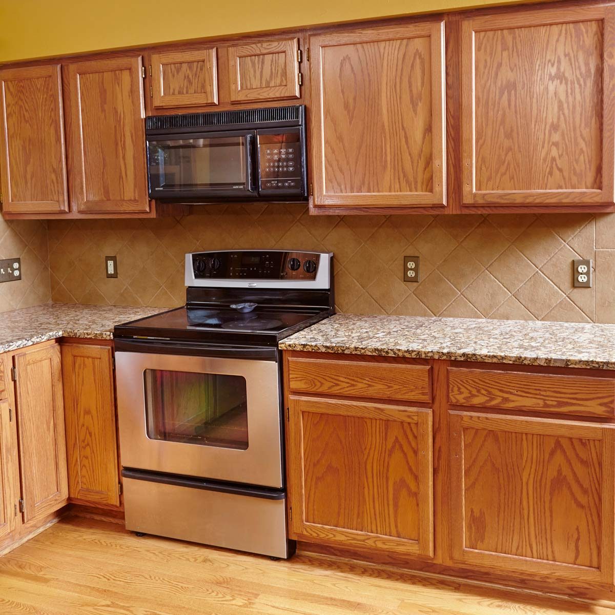 A stainless steel stove stands in a kitchen, surrounded by wooden cabinets and granite countertops, with a microwave mounted above and tile backsplash behind.