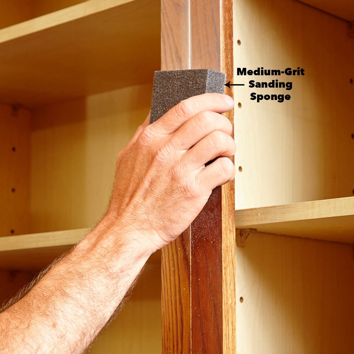 A hand with a medium-grit sanding sponge smooths a wooden edge in a bright, uncluttered shelving area, enhancing the wood's finish.