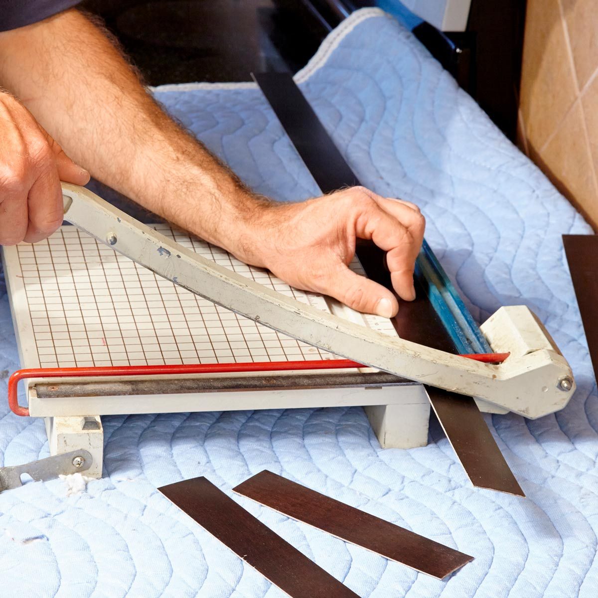 A hand operates a paper cutter, slicing through sheets, on a blue padded surface. Several cut strips lie scattered nearby.