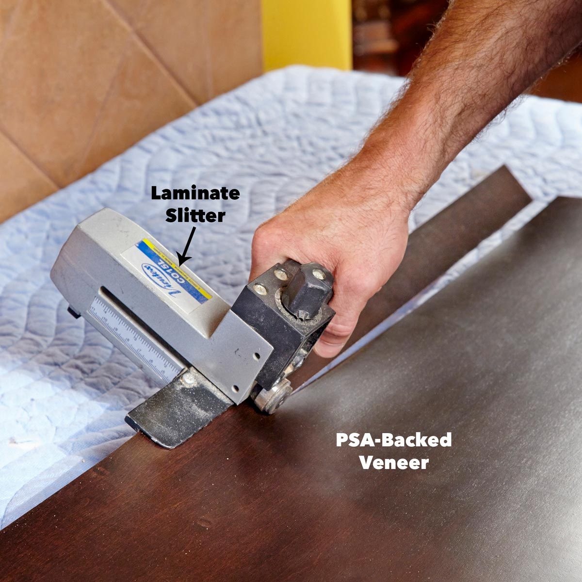 A hand holds a laminate slitter cutting through a dark, smooth veneer on a quilted surface, indicating a woodworking or crafting activity.