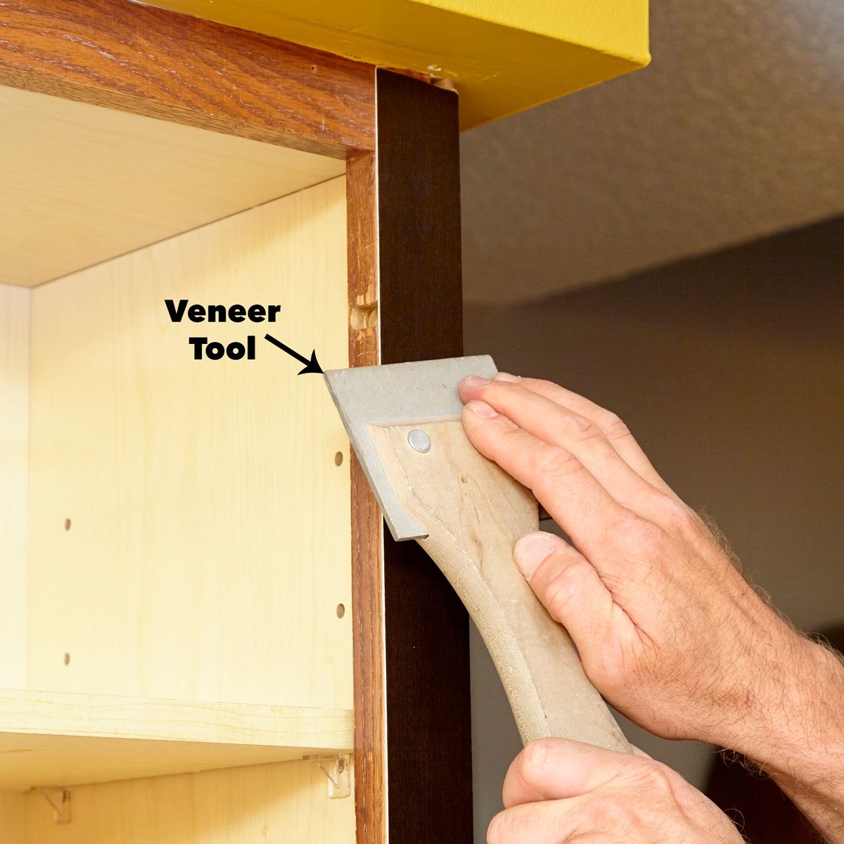 A person uses a veneer tool to scrape the edge of a wooden cabinet in a well-lit interior space, focusing on detailing.