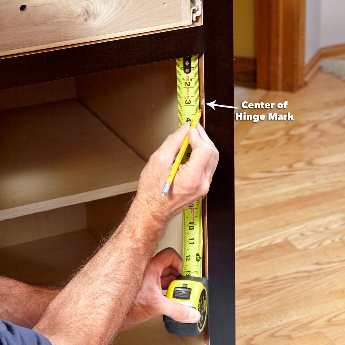A hand holds a pencil while measuring a marked point on a wooden cabinet with a tape measure, surrounded by a wooden floor and interior space.