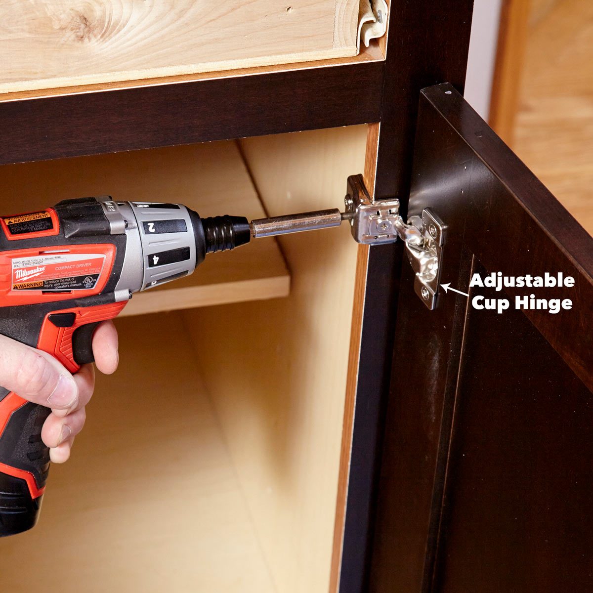 A power drill is tightening a cup hinge onto a cabinet door, inside a wooden cabinet with a shelf visible in the background.