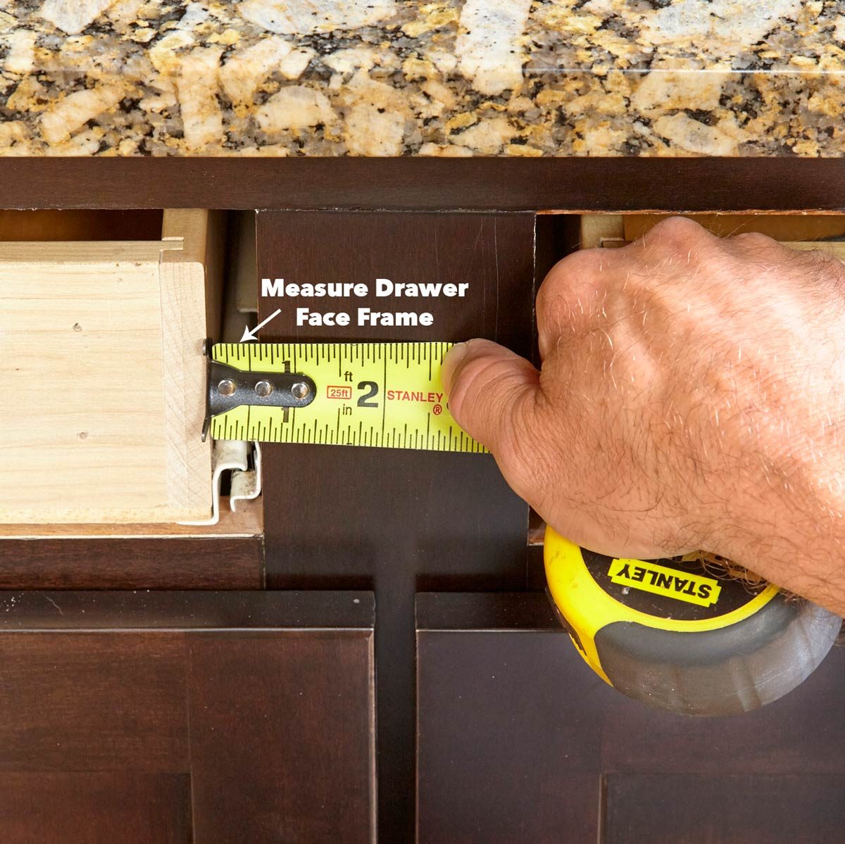 A hand holds a tape measure against a drawer's face frame, measuring its dimensions near a granite countertop. The environment suggests home improvement or carpentry.