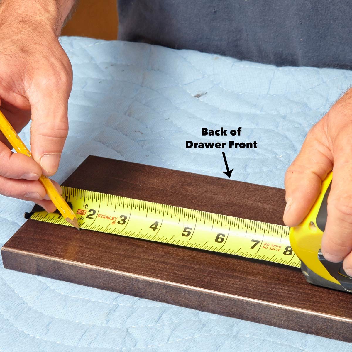 A hand holds a pencil marking a wooden board, while another hand measures it with a yellow tape measure, on a blue padded surface.
