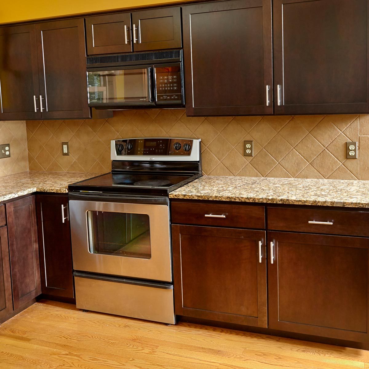 Kitchen cabinets with a stainless steel oven and microwave prepare in a well-lit space featuring granite countertops and a tiled backsplash against wooden flooring.
