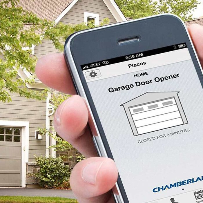 A hand holds a smartphone displaying a garage door app, indicating the door is closed for three minutes, against a house backdrop with greenery.