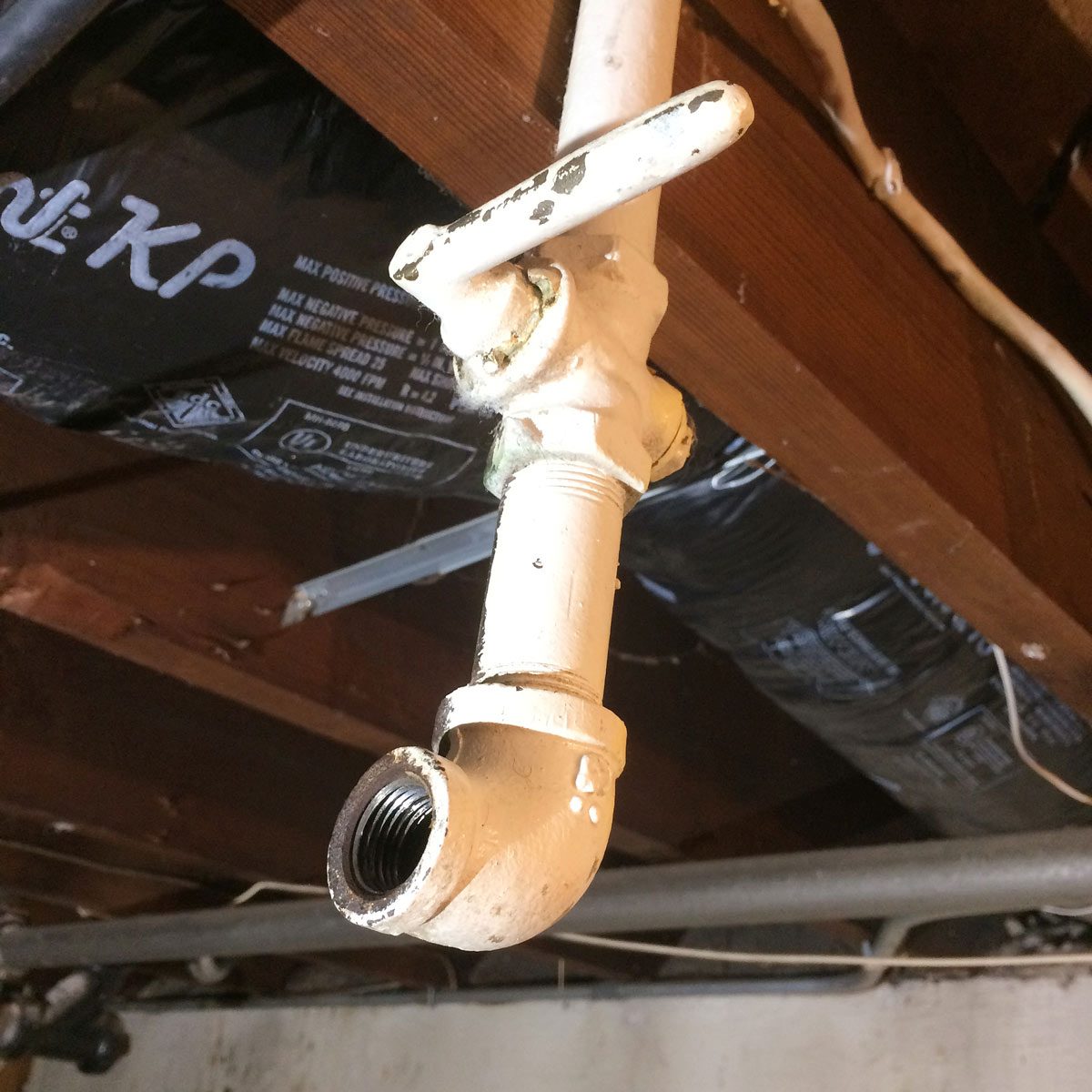 A white pipe fitting with a valve hangs from a wooden structure, leading to an elbow joint with threading in a dimly lit basement environment.