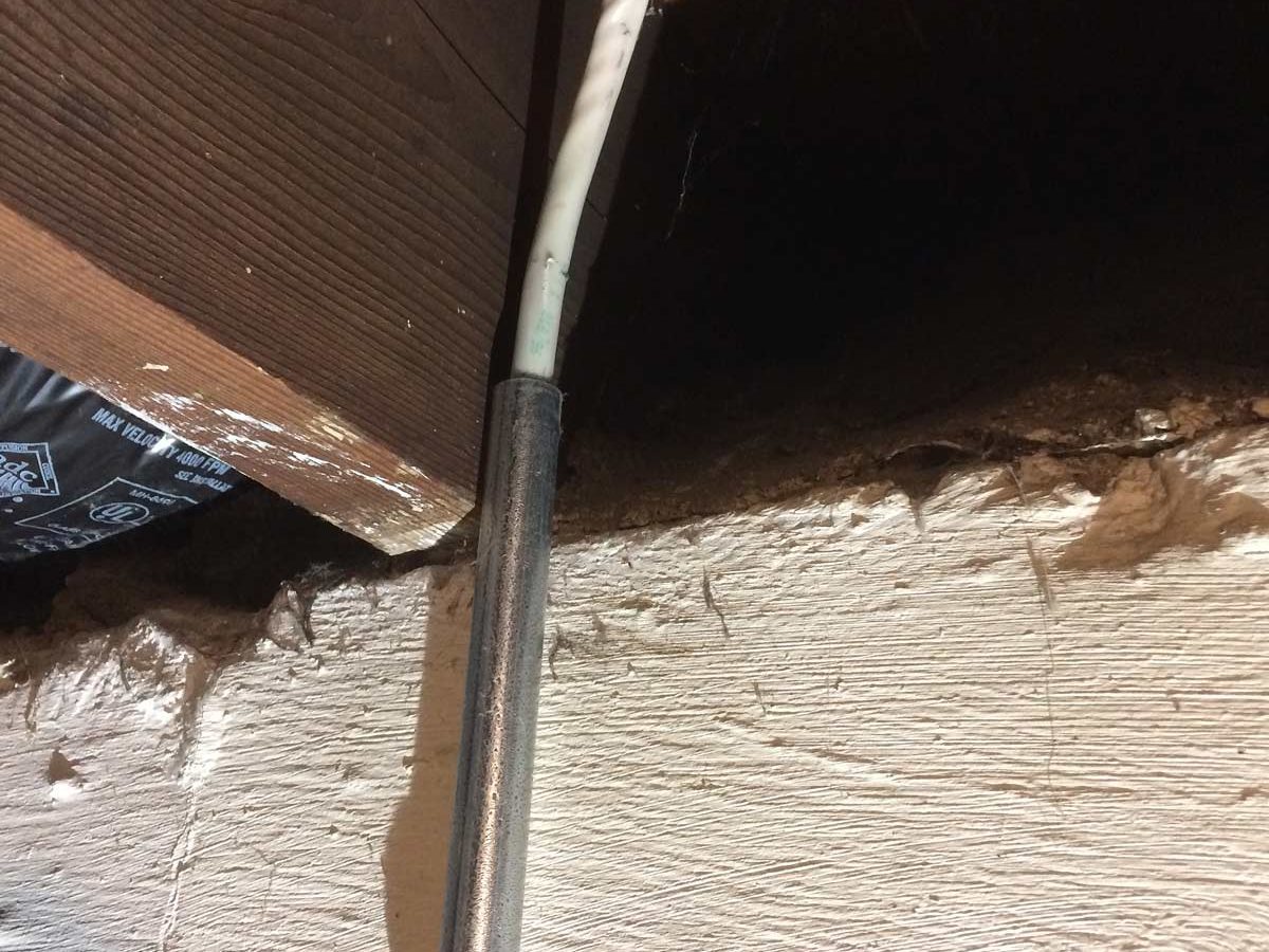 A metal pipe runs vertically along a wall, connecting to white electrical wiring beneath a wooden beam in a dimly lit basement space.