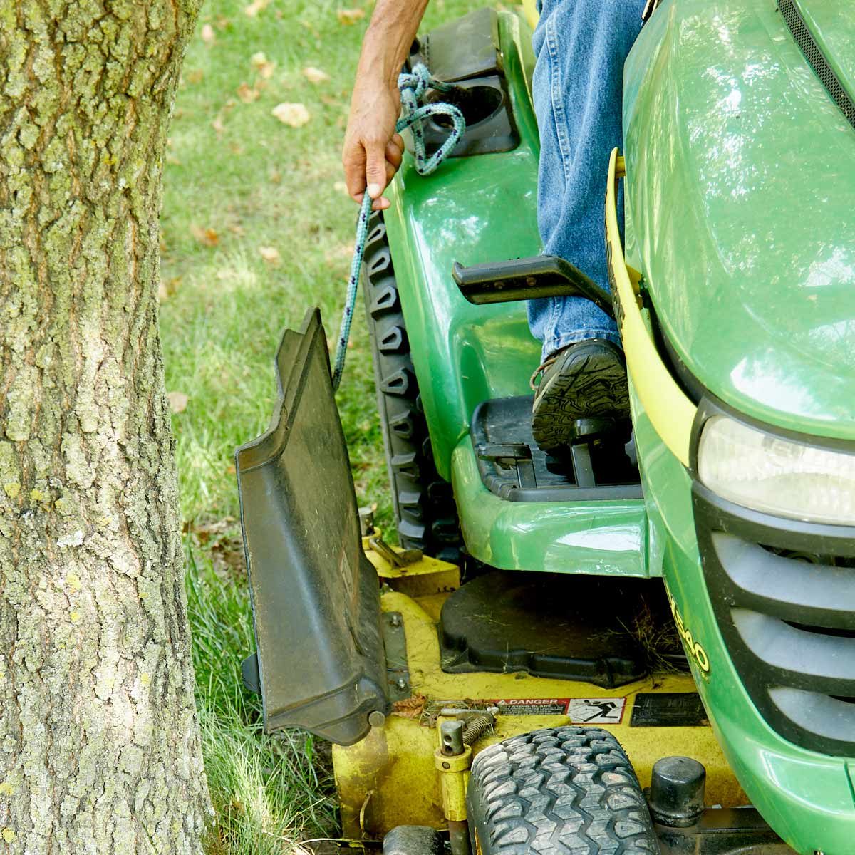 A person pulls a rope while sitting on a green lawn mower, positioned near a tree in a grassy yard.