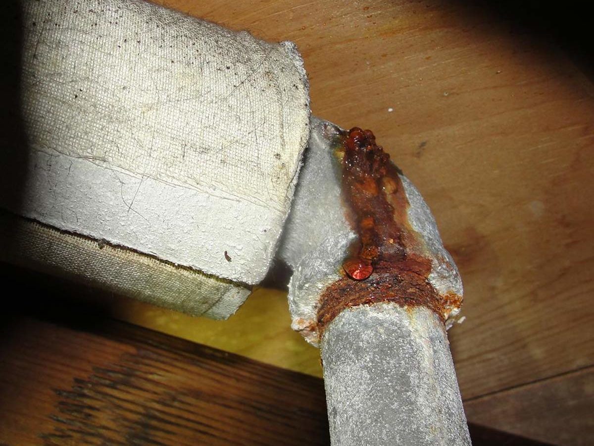 A corroded metal pipe joint leaks water onto a wooden surface, surrounded by a textured fabric covering and wooden beams in a dimly lit area.