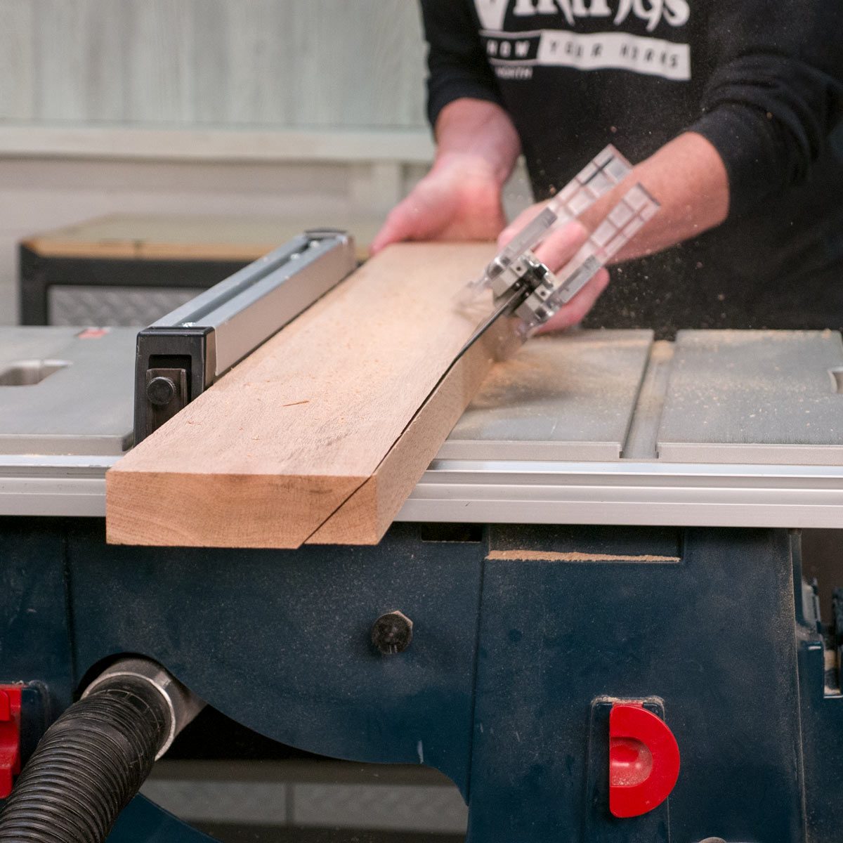 A person feeds a wooden plank through a table saw, creating sawdust in a workshop environment. The machine supports precision cutting.