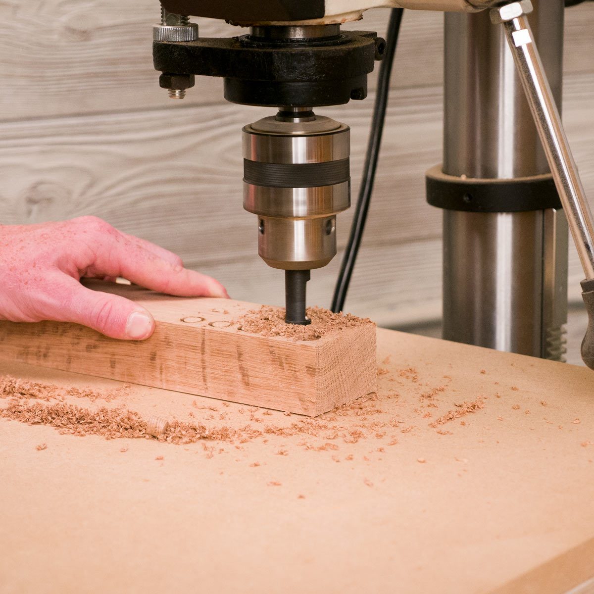 A drill bit is descending into a wooden block, creating shavings, while a hand holds the block against a work surface in a workshop.