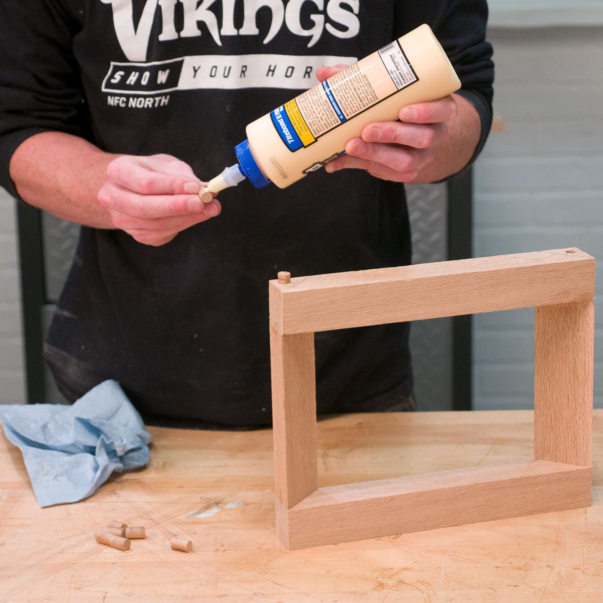 A person applies wood glue onto a wooden dowel, preparing to join it with a wooden frame on a workbench, surrounded by tools and materials.