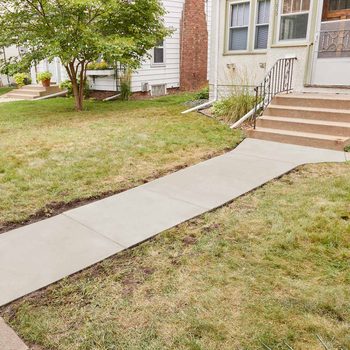 A concrete walkway extends from a grassy area toward a set of stairs leading to a house, surrounded by a mix of grass and plants.