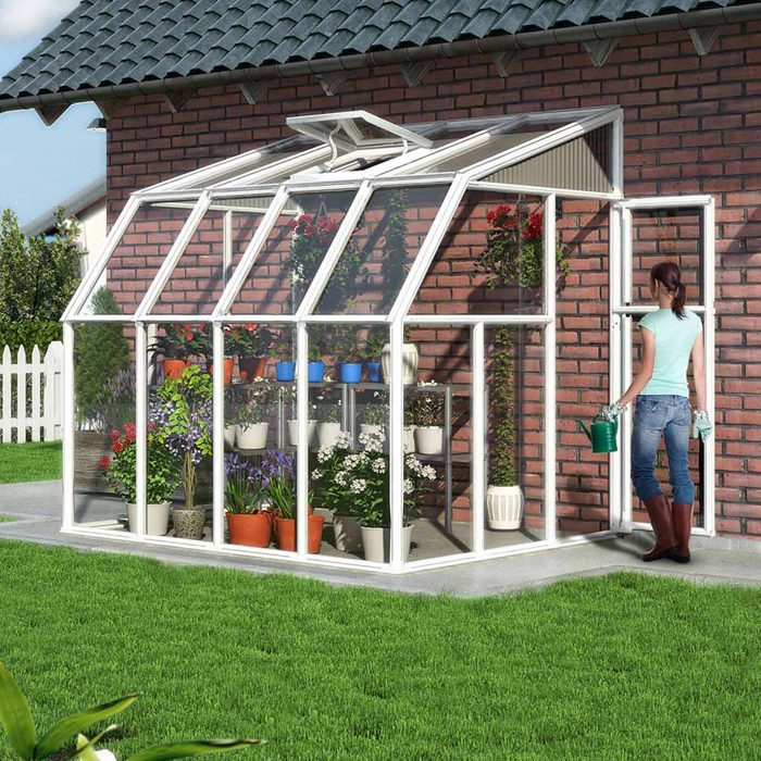 A woman with a watering can tends to plants inside a glass greenhouse beside a brick house, surrounded by a grassy lawn and white fence.