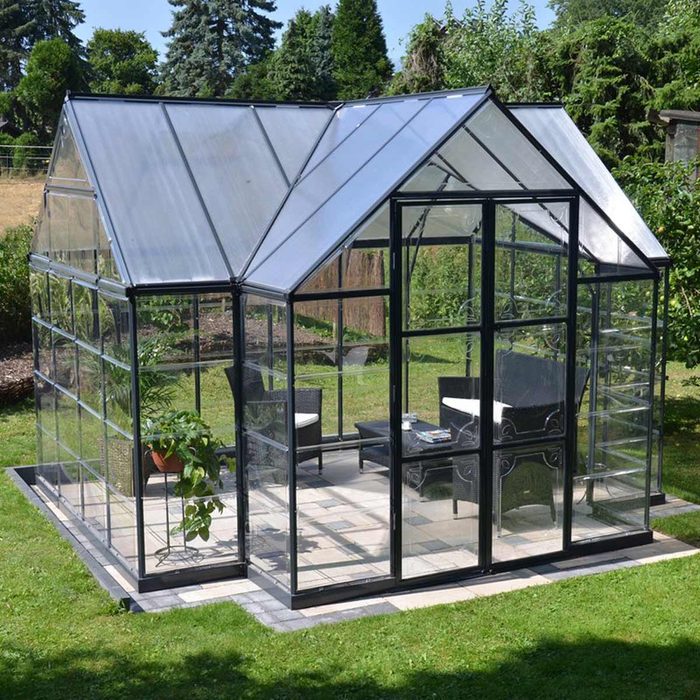 A greenhouse features transparent walls and roof, housing a seating area with a table, surrounded by greenery in a sunny garden setting.