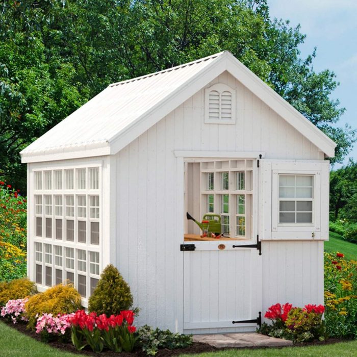 A small white shed stands among colorful flowers, with an open window displaying gardening tools, surrounded by lush greenery under a clear blue sky.