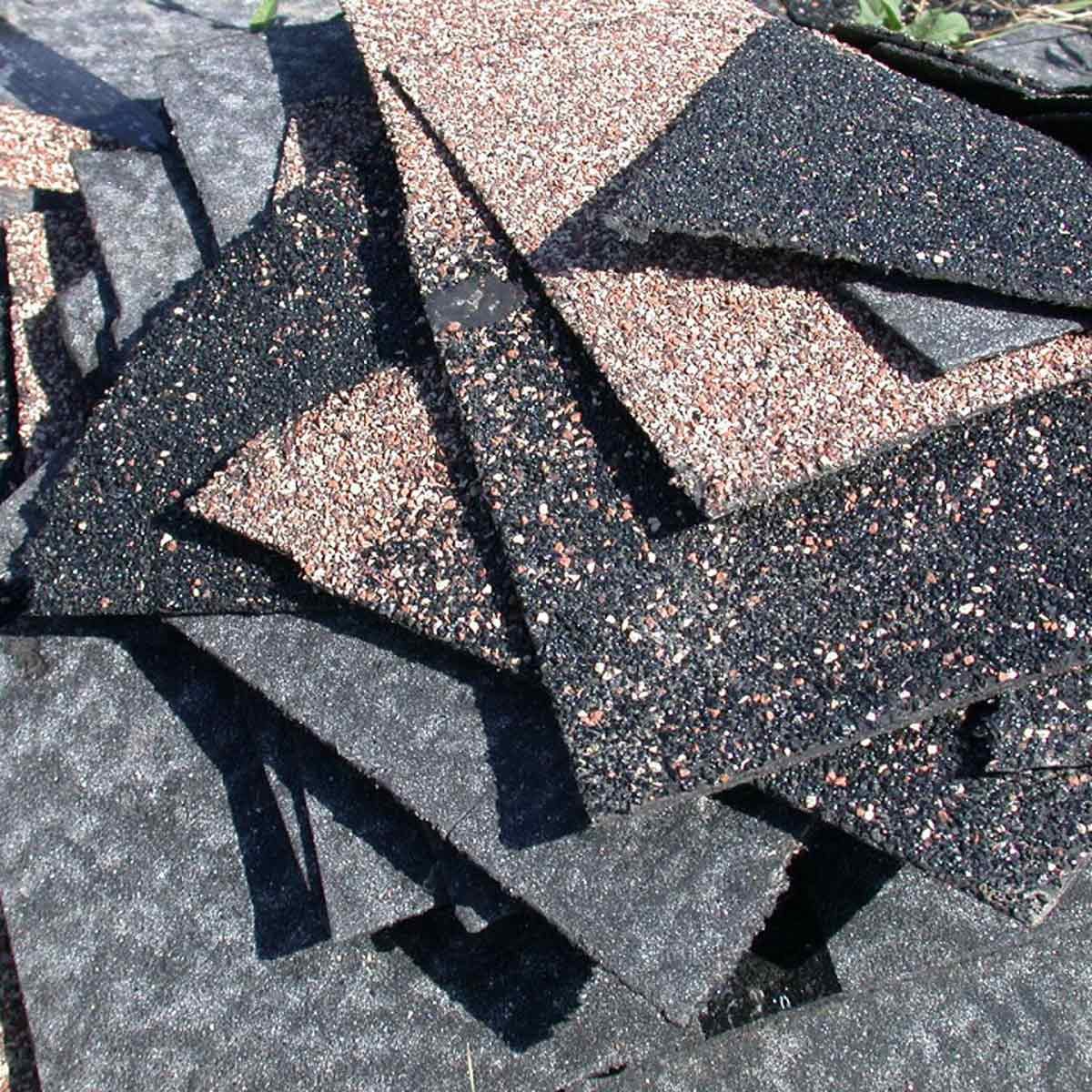Several asphalt shingles are stacked in disarray, showing varying textures and colors, on a surface outdoors, likely awaiting installation or disposal.