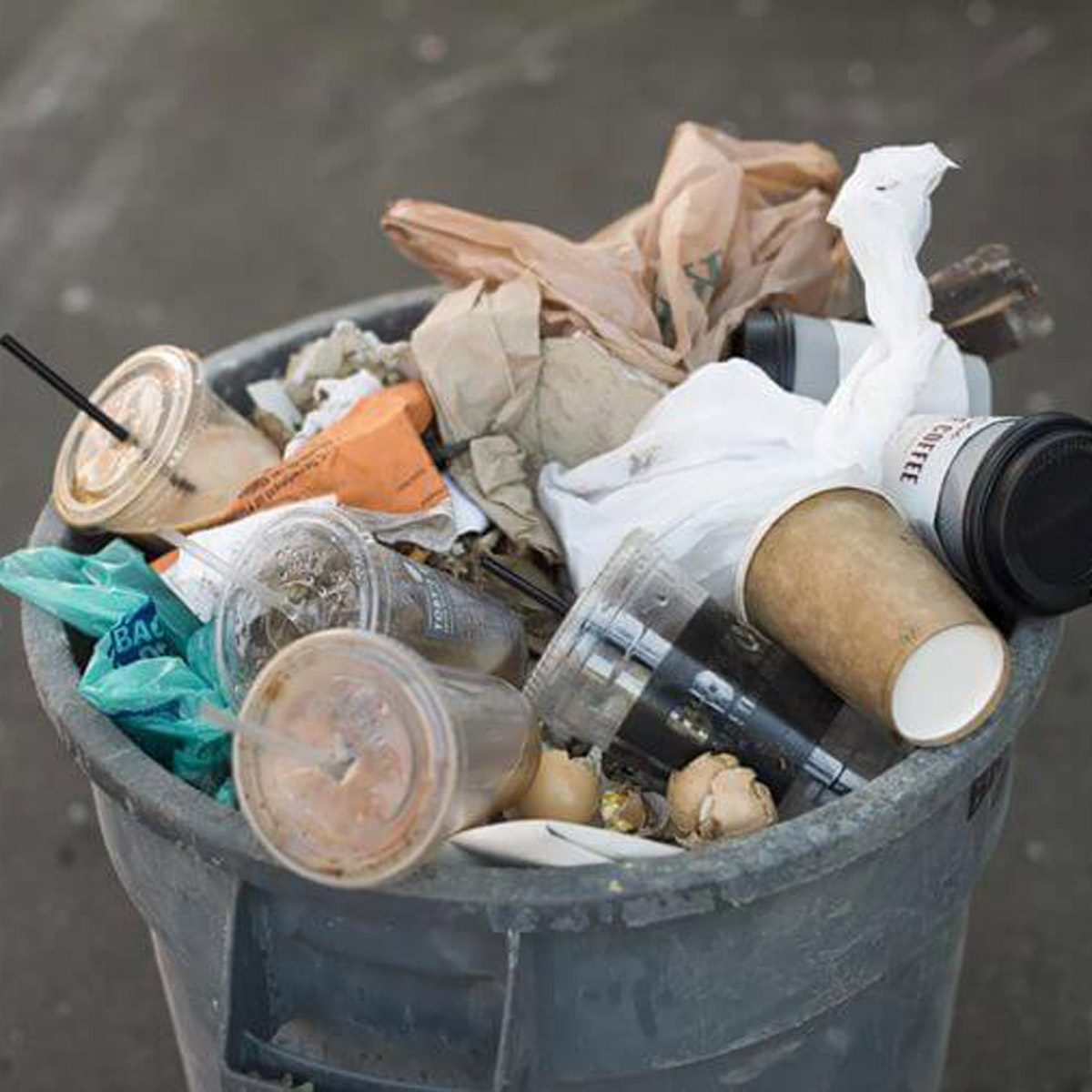 A gray trash can overflows with various discarded items, including food wrappers, plastic bags, and coffee cups, indicating an untidy environment.