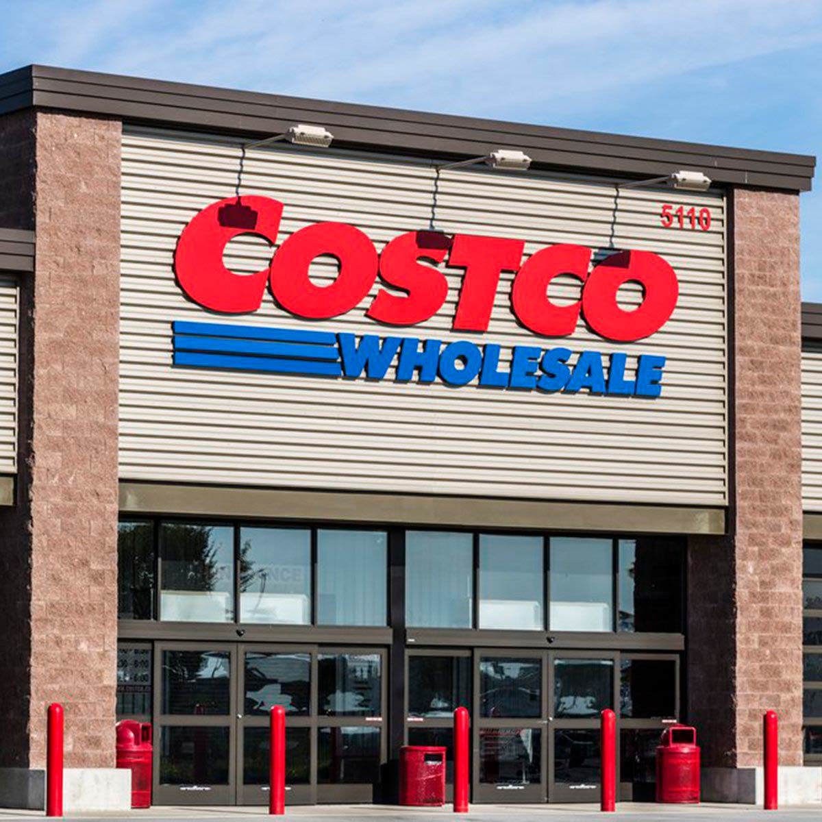 A large retail building displays a bright red sign with "COSTCO WHOLESALE," while red poles and shopping carts are positioned outside under a clear blue sky.