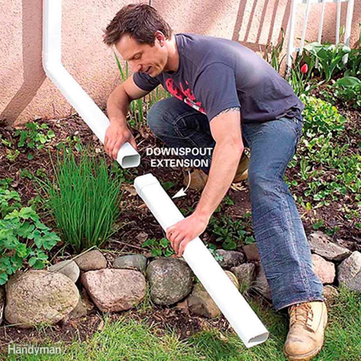 A man connects a white downspout extension to a gutter. He is kneeling in a garden with rocks and plants surrounding him.